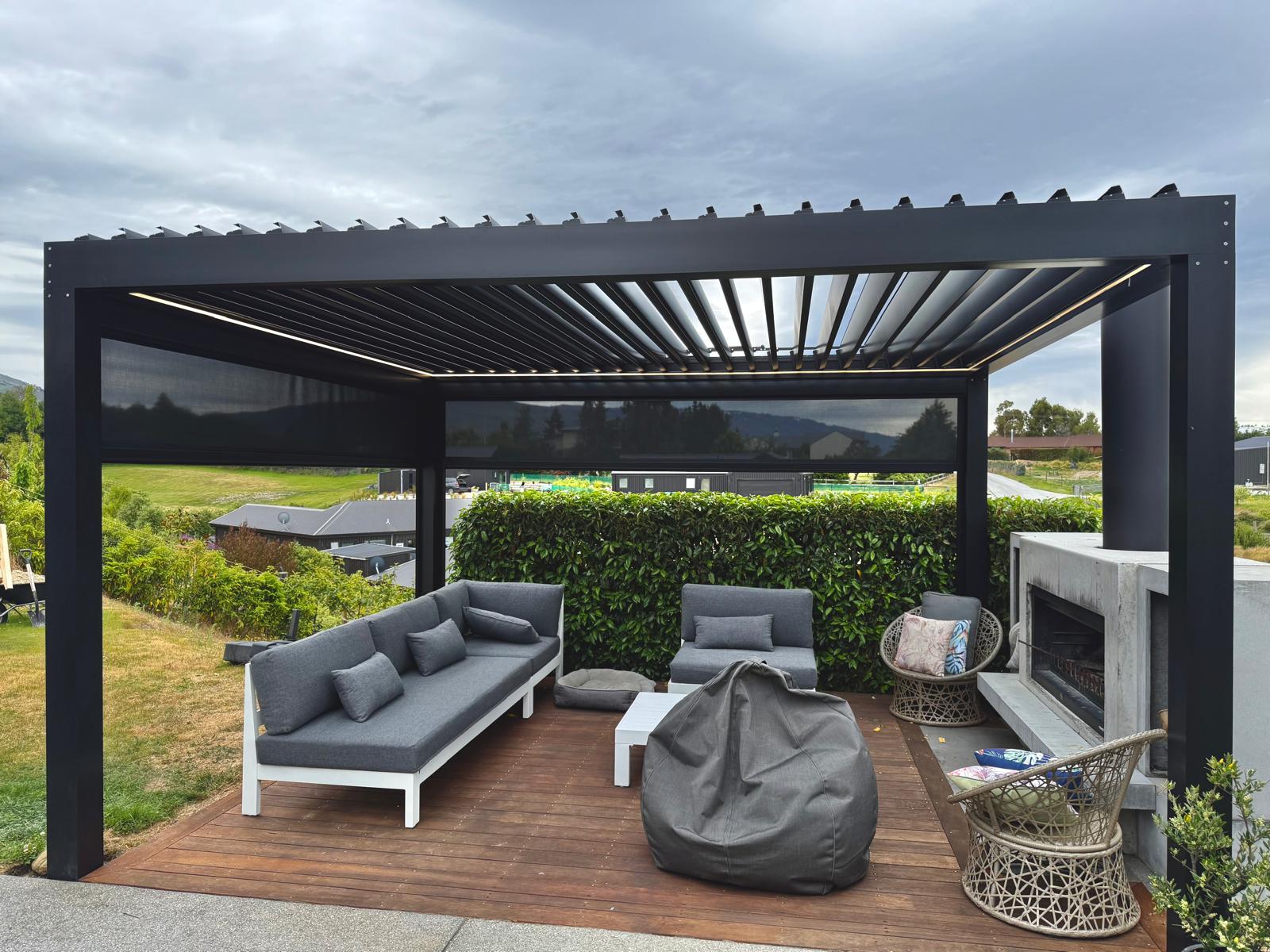Black louvre roof with LED lighting and outdoor blinds over a deck and concrete fireplace in Central Otago